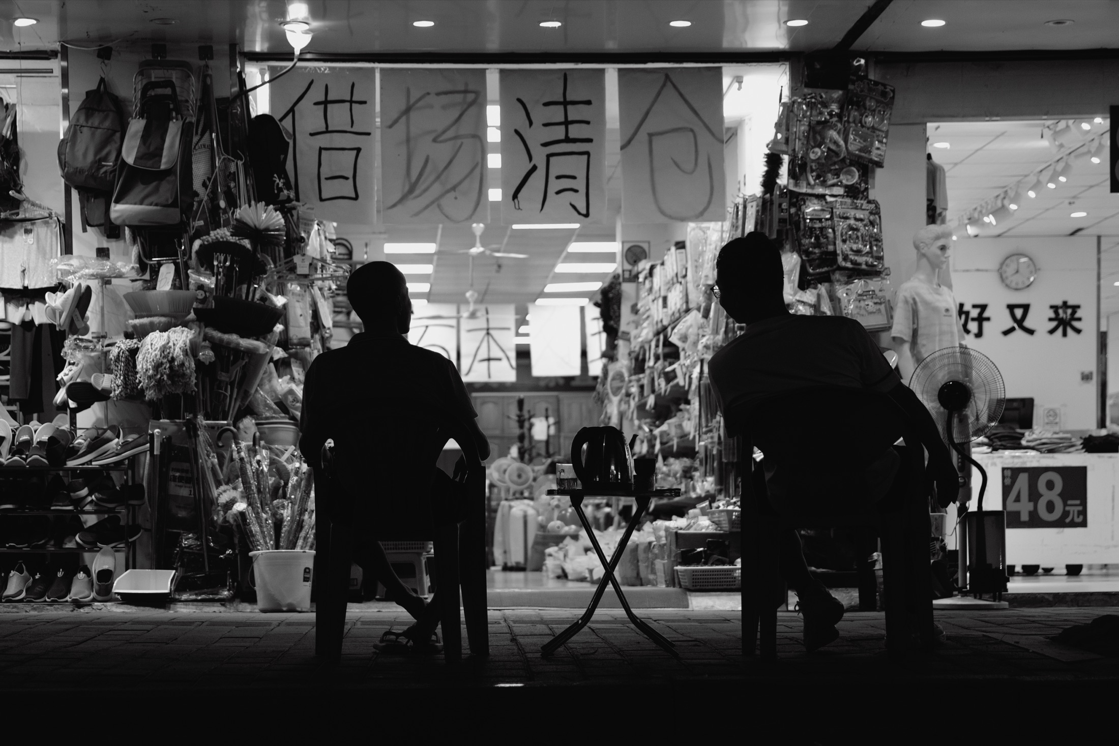 黑白夜景中，两个人坐在亮着灯的杂货店门前